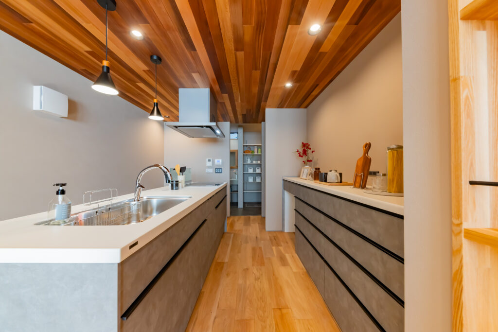 レッドシダーの板張り天井が印象的な、グレーの腰壁がおしゃれな対面式オープンキッチン。背面のカップボードも同系色で統一し、無垢の床材やペンダントライトが調和する、モダンで温かみのあるキッチンデザインです。
