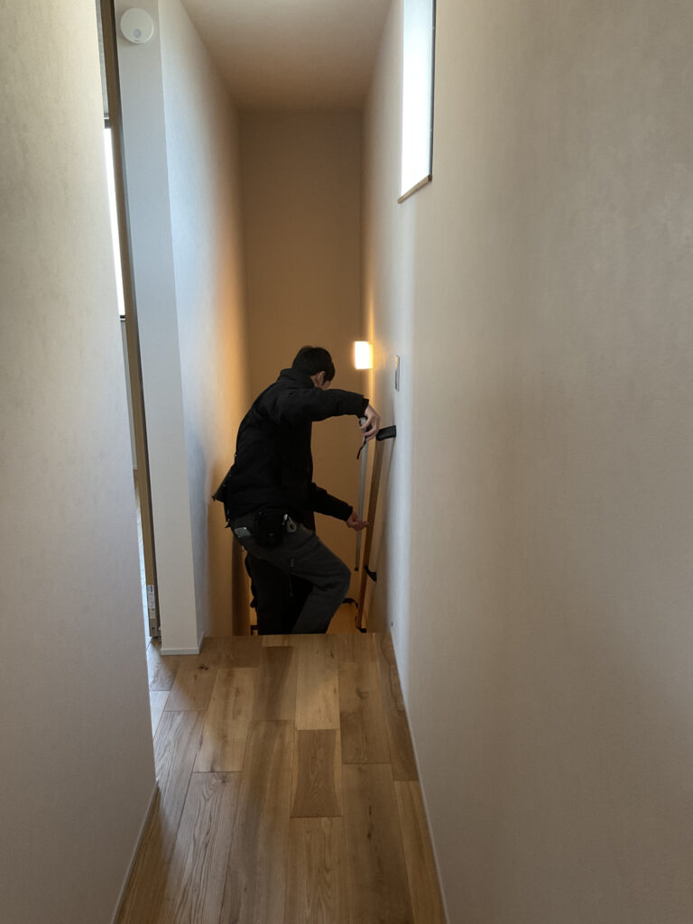 新築住宅の完成時検査で、階段の手すりを取り付ける職人の様子。木目調のフローリングと白い壁の廊下。