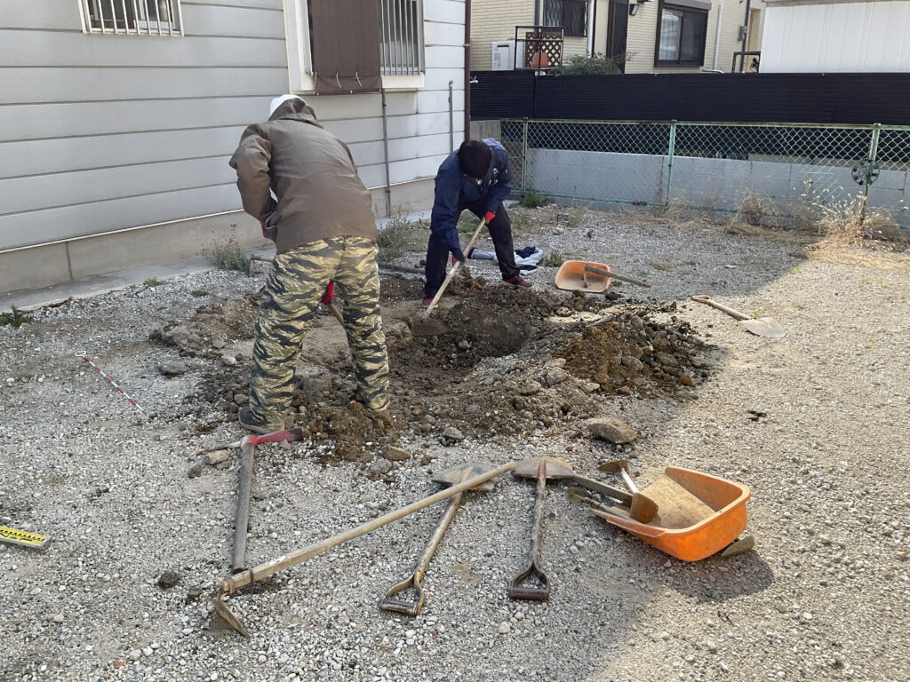 更地において、スコップやツルハシなどの手道具を使用して行われている埋蔵文化財の試掘調査。作業服のスタッフが地面を丁寧に掘り進めている現場風景。