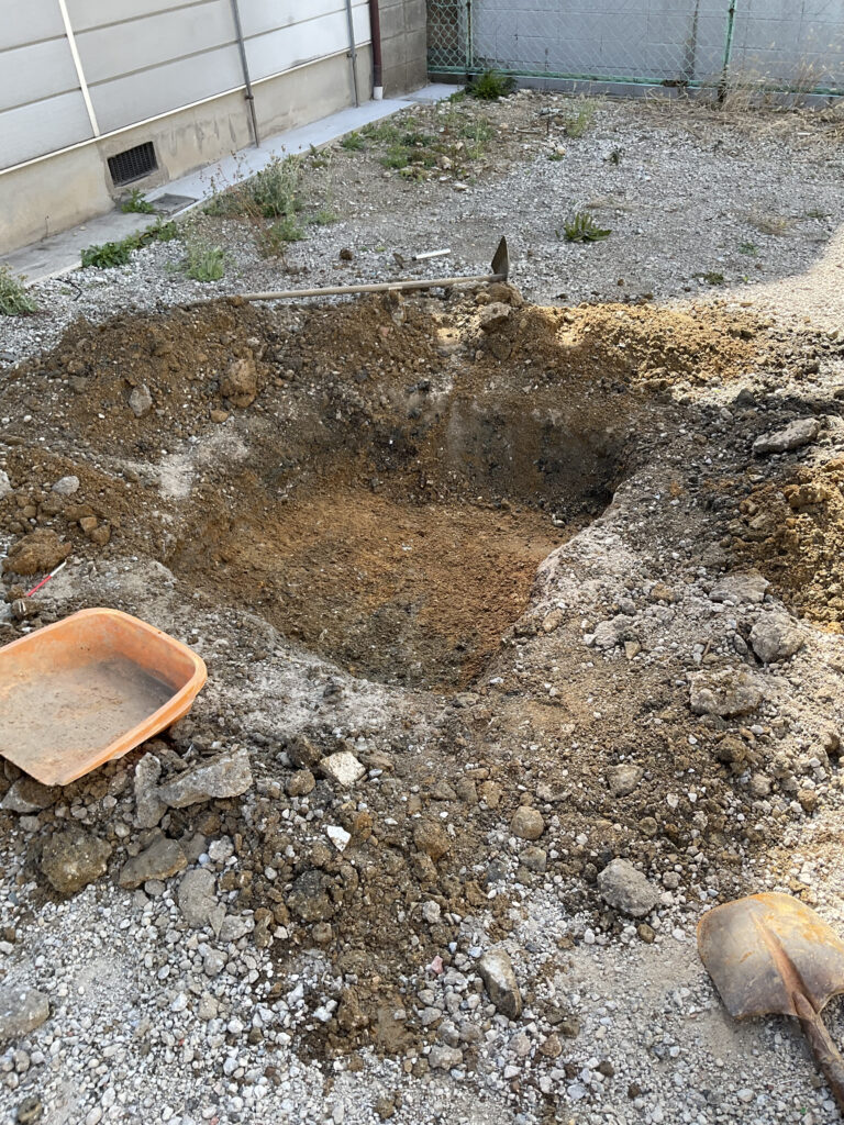 地中の遺構を確認するために掘削された試掘坑（トレンチ）。掘り起こされた土に囲まれ、土層の重なりを確認できる四角い掘削穴と、周囲に置かれた調査道具。