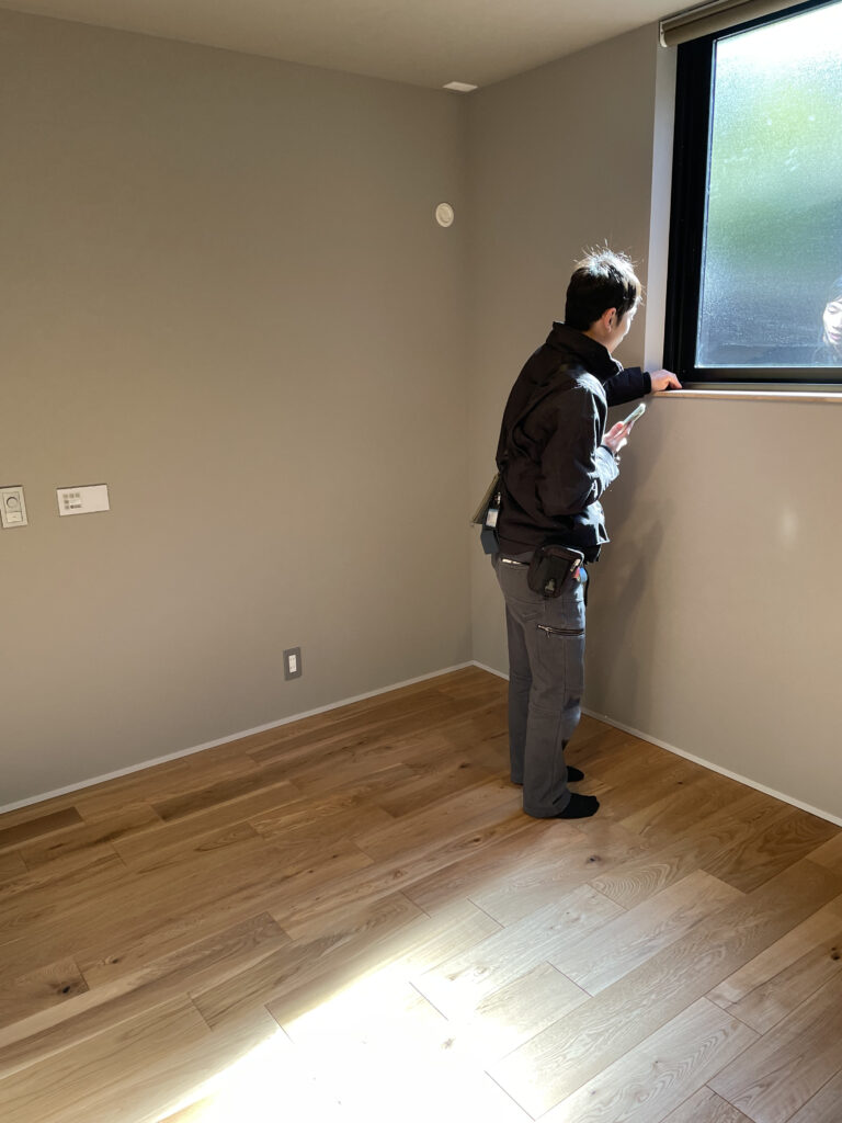 完成前の住宅検査。グレーの壁の部屋で窓枠の状態や眺望をチェックする検査員の男性。