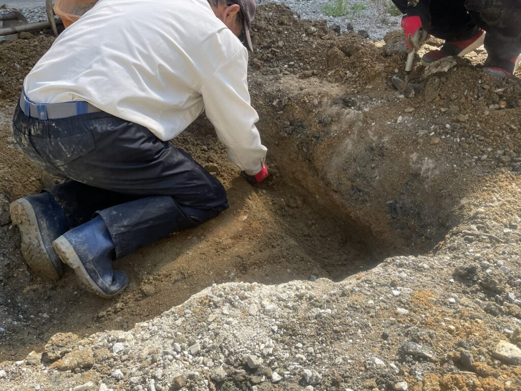 掘削された穴の中で、手道具を使い地層を詳細に確認する作業員。建築予定地の地下に歴史的な価値があるものが残っていないかを判断するための精密な調査。