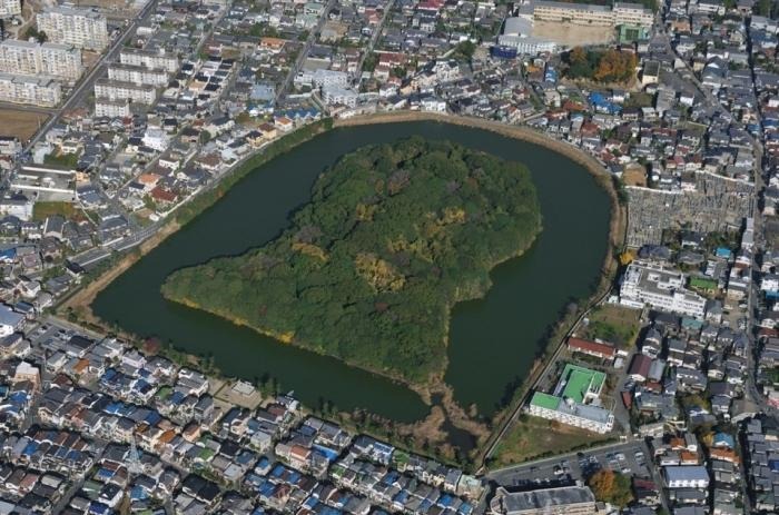 住宅街の中に位置する、深い緑に覆われた前方後円墳の全景。周囲を囲む濠（ほり）と、歴史的遺構を守るように建ち並ぶ現代の家々が対照的な空撮写真。
