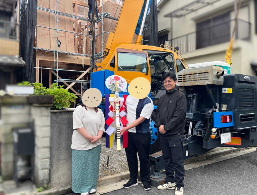 建築現場のクレーン車の前にて、上棟式で使われる御幣(ごへい)を大切そうに持つご夫婦と、笑顔で並ぶ工務店スタッフの記念写真。