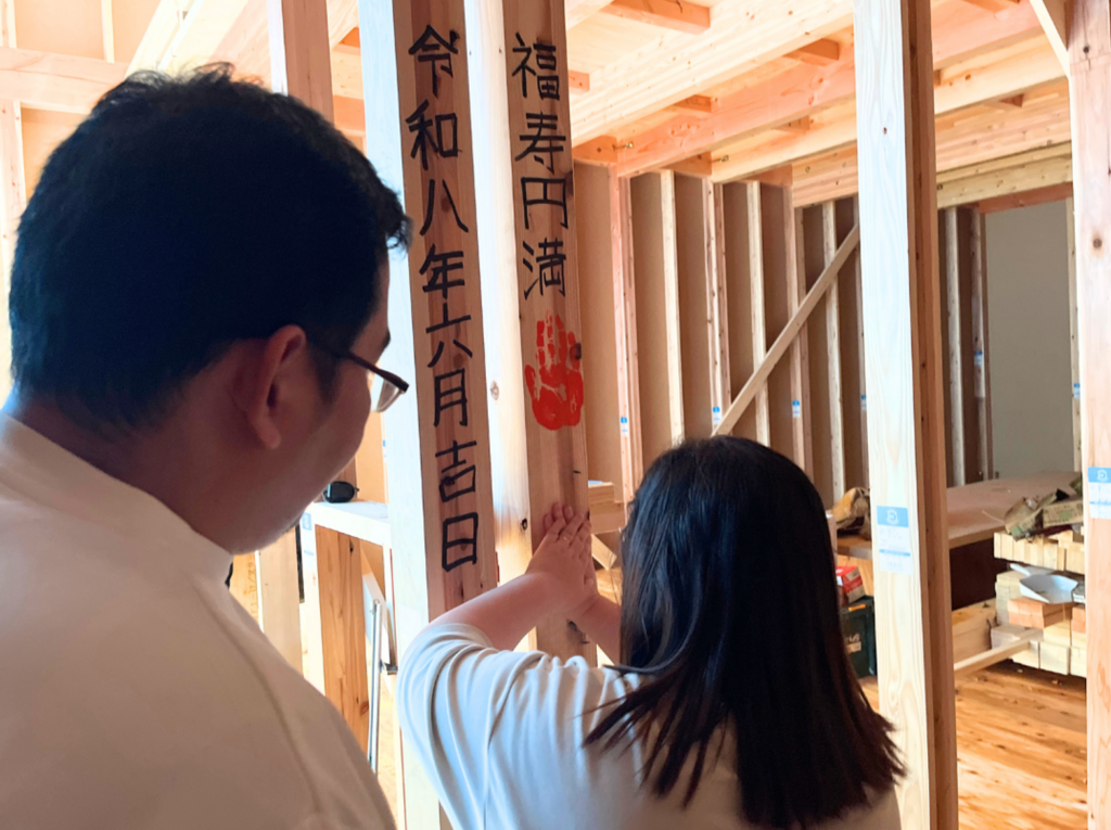 「令和八年六月吉日」「福寿円満」と記された構造柱に、記念の赤い手形を真剣な表情で慎重に押し当てているご夫婦の後ろ姿。