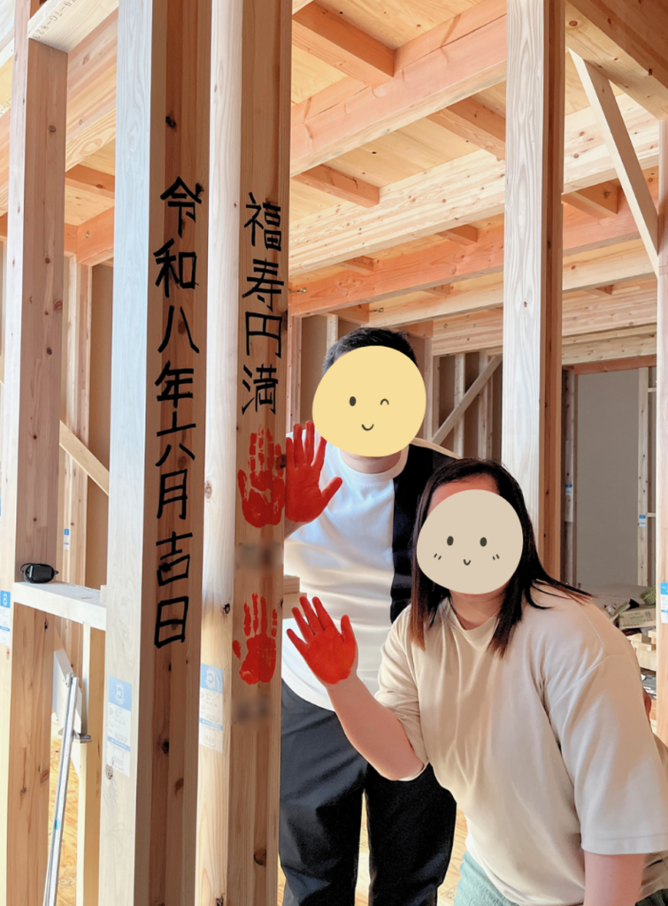 建築中の構造躯体の中で、墨文字で「令和八年六月吉日」「福寿円満」と書かれた大黒柱に、赤い手形を押し終えて笑顔を見せるご夫婦。
